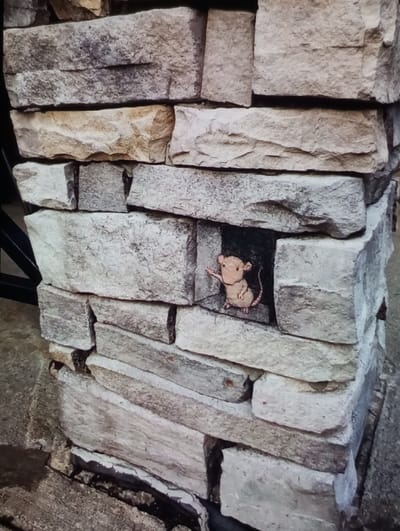The image displays a section of a stone pillar or wall located in Aktash, Uzbekistan. The structure is composed of irregularly shaped, rough-hewn stone blocks in various shades of grey, beige, and light brown, stacked with visible mortar lines. Approximately centered vertically on one face of the structure, a dark, rectangular recess or niche is present between two larger stones. Within this niche, a painted depiction of a light brown mouse is visible. The mouse, rendered in a cartoon style, exhibits pink ears, nose, and paws, dark eyes, and a black tail extending to the right. Its left paw is raised in an upward and slightly forward gesture, implying it is emerging or gesturing from the opening. The base of the stone structure rests on a grey concrete or paved surface, which shows minor cracks and dark debris in the crevices where it meets the ground. A dark, angular object, possibly a metallic support or railing, is partially visible in the background to the left of the stone structure.