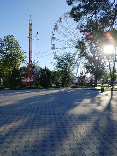 This is a bright, sunny daytime photograph of an amusement park in Vinogradny, Russia. In the background, a large white Ferris wheel is visible against a clear blue sky. To the left of the Ferris wheel, a tall, red amusement ride with a pointed top extends upwards. There is a swing ride with people seated on it beneath the tall red structure. Trees with green foliage surround the rides and extend into the foreground on the right side of the image. The ground in the foreground is paved with light gray interlocking rectangular stones, with a patterned shadow cast across it from the trees and possibly the rides. A lamppost stands between the Ferris wheel and the tall red ride. In the middle ground, a pathway with people walking can be seen, and further back, more trees and structures are present. The sun is shining brightly, creating lens flares in the upper right portion of the image, and casting long shadows.