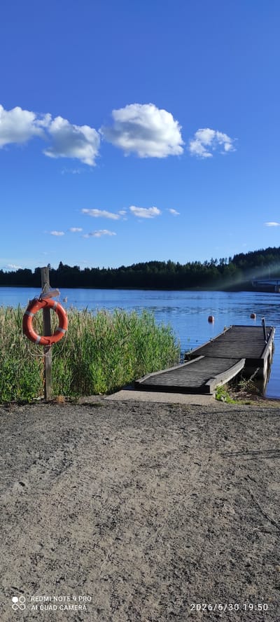 The image depicts a serene lakeside scene in Joutsa, Finland, bathed in the warm glow of late afternoon sunlight. The foreground is dominated by a sandy and gravelly path, showing signs of foot traffic. To the left, a weathered wooden post supports a bright orange lifebuoy. Beyond this, tall green reeds line the water's edge. A wooden pier extends into the calm, blue water, with a sloping ramp providing access. Two orange buoys float near the pier. The lake stretches towards a dense, dark green forest that forms the horizon, its silhouette sharp against the vibrant blue sky dotted with fluffy white clouds. In the distance, a bridge can be seen crossing the water. The overall atmosphere is one of tranquility and natural beauty, suggesting a popular spot for outdoor recreation in Joutsa.