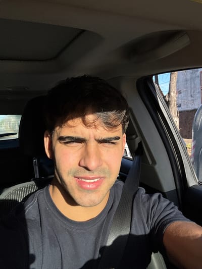 In Gerli, Argentina, a young man is captured in a selfie taken from inside a vehicle. He is seated in the driver's position, with a seatbelt fastened across his dark t-shirt. The sunlight streams in from the right, casting a strong shadow across the left side of his face and body. The interior of the car is dimly lit, contrasting with the bright exterior visible through the open window. Outside, a tree and a building are partially visible, suggesting an urban or suburban setting. The man's expression is neutral, with his gaze directed towards the camera.
