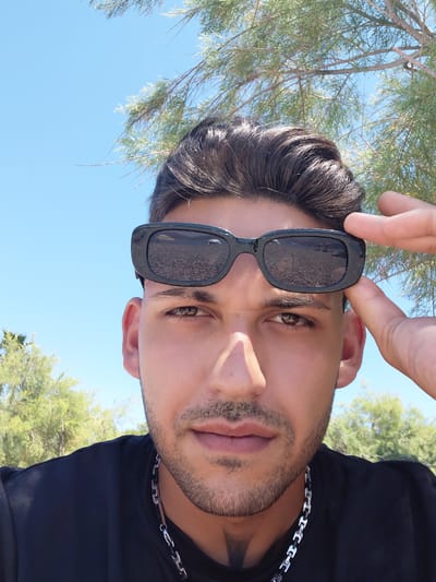 The image is a close-up, self-taken photograph of a young man wearing sunglasses and a black t-shirt, with a chain necklace. He is outdoors, with trees and a clear blue sky in the background. The sunglasses reflect a pebbled beach with some figures visible. The lighting suggests it is daytime. The location is identified as the Municipal Unit of Aegio, Greece.