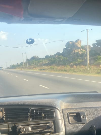 The image depicts a view from inside a vehicle traveling on a paved road in Unguwar Fatika, Nigeria. The landscape outside the vehicle consists of rocky formations and sparse vegetation. A utility pole is visible along the roadside. Inside the vehicle, a dashboard with an air vent and a locking mechanism are visible. A Sony device is also present on the dashboard. There are no people visible in the image.