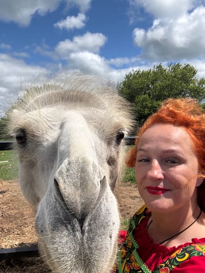 A close-up, slightly upward-angled selfie features a woman with vibrant orange hair and a red, ornately patterned dress, smiling directly at the camera. Beside her, dominating the left side of the frame, is the head of a light-colored camel, its large, dark eye visible. The background consists of a bright blue sky with scattered white clouds, a dense green tree line, and a dirt enclosure with scattered straw. The woman is wearing red lipstick and has noticeable eye makeup. The camel appears to be facing the camera, with its snout close to the lens. The overall impression is a candid, outdoor snapshot taken on a sunny day. The location is specified as the city of Tri Ozera, Russia.