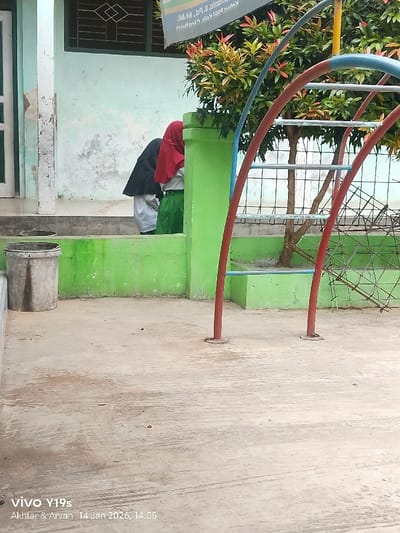 Two girls wearing hijabs stand near a green painted wall. One wears a black hijab, the other a red one. A climbing frame painted red and blue is nearby. A bush is growing behind the climbing frame. A bucket sits near the wall. The ground is concrete. A sign hangs above, attached to the building's exterior.