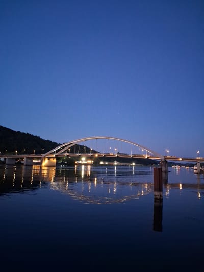 A long shot captures the reflection of a bridge and lights on a body of water at dusk in Vilshofen an der Donau, Germany. The sky is a deep blue, transitioning to black towards the top of the frame. A modern, arched bridge spans the width of the image, illuminated by numerous lights along its structure and on the roadbed. The lights from the bridge and distant structures create a shimmering reflection on the dark, calm water. On the left side of the frame, a dark, tree-covered hillside rises behind the bridge. The right side of the water features a dark, vertical post and further reflections of lights from what appear to be boats or buildings. The scene is serene and atmospheric, highlighting the interplay of artificial light and the natural environment.