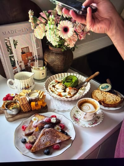A flat lay image depicts an elaborate breakfast or brunch spread laid out on a light-colored surface. A person's hand is visible on the left side, holding a smartphone as if taking a photograph or video of the arrangement.

The meal features several dishes: French toast slices dusted with powdered sugar, garnished with fresh raspberries, blueberries, and small white daisies, presented on a white plate with a gold rim. Another plate holds toasted bread slices, possibly sourdough, accompanied by a small round tin with a partially visible label "HENRY." A scallop-edged bowl contains what appears to be a creamy dip, such as whipped feta or yogurt, topped with sliced avocado, dill, and paprika, with a small knife resting in it. A wooden serving board displays an assortment of crackers, cheese cubes, and a small container of spread, also adorned with fresh raspberries. Beverages include a coffee with distinct latte art (a leaf design) in a floral cup and matching saucer, and an empty floral mug.

Complementing the food is a bouquet of pink gerbera daisies and other light-colored flowers arranged in a rustic vase. A large hardcover book titled "MADE FOR LIVING" is visible, along with a candle in a glass jar, which appears to be a Jo Malone brand candle. The scene conveys a sense of indulgence and aesthetic pleasure, typical of a leisurely indoor breakfast or brunch. No specific location cues for Ros Na Rí, Ireland are visible within the image itself.