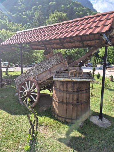 The image depicts a rustic wooden barrel and an old-fashioned cart situated on a grassy area. A red-tiled roofed structure, likely a shelter, partially covers the barrel and cart. The barrel is large and made of wooden staves banded with metal hoops. The cart is intricately constructed with woven sides and a large wooden wheel with spokes. A basket is visible hanging from the cart. In the background, lush green trees and a mountain covered in foliage can be seen, suggesting a natural, perhaps rural, setting. A paved area with vehicles is visible in the distance, implying proximity to a road or parking area. The scene is bathed in natural light, suggesting it is daytime, possibly with sunny weather, as indicated by a rainbow-like light flare in the lower right portion of the image. The overall impression is one of traditional craftsmanship and a peaceful, outdoor environment. There is no visible text in the image.