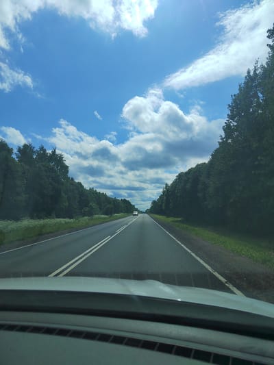 The image shows a view from inside a vehicle looking out at a two-lane asphalt road lined with dense green trees on both sides. The sky above is a bright blue with scattered white cumulus clouds. A white car is visible in the distance on the road. The foreground shows the dashboard and windshield of the vehicle. The scene appears to be a rural or suburban area, possibly on the outskirts of Vitino, Russia, given the presence of trees and a relatively open road.