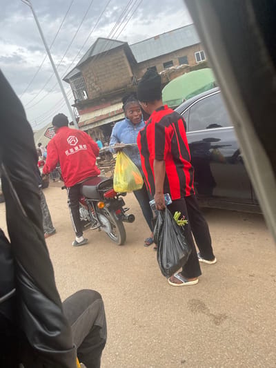 In Gwong, Nigeria, a street scene unfolds under an overcast sky. A motorcyclist, wearing a red jacket with white Chinese characters and black pants, is stopped. Beside him, a woman in a light blue denim jacket and dark pants holds a plate of food and a yellow plastic bag. To her right, another person in a red and black striped shirt and black pants holds a black plastic bag containing green vegetables and a smartphone. A black car is parked behind them. The background shows a brick building with a tin roof, suggesting a commercial or residential area. The ground is paved. The image appears to be taken from a moving vehicle, possibly a trishaw, due to the blurred foreground elements.