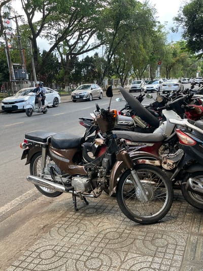 The image depicts a street scene in Liên Nghĩa, Vietnam, with a focus on several parked motorcycles and scooters on a sidewalk adjacent to a road. A brown Honda Super Cub motorcycle is prominently in the foreground, parked on a patterned tiled sidewalk. Behind it, a variety of other motorcycles and scooters are tightly packed together. In the background, a white car with license plate "43B-251.21" is visible on the asphalt road, with a scooter carrying two people on it further ahead. Several other cars, including silver and white sedans, are also on the road. The environment includes trees lining the street and overhead power lines, suggesting an urban or semi-urban setting. The lighting indicates it is daytime, likely overcast.