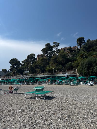 The image depicts a pebble beach in Finale Ligure, Italy, under a bright blue sky with scattered white clouds. The foreground is dominated by the textured grey pebbles of the beach. In the mid-ground, rows of unoccupied green sun loungers and matching umbrellas are neatly arranged. Some people are visible lounging on sunbeds, one woman is lying down on a sun lounger in the left mid-ground, and others are walking or standing in the distance near a row of buildings. These buildings, situated behind the loungers, are multi-story structures with balconies and windows, some painted in warm earth tones. Lush green trees and vegetation cover the hillside behind the buildings, with taller pine trees and palm trees interspersed. The overall scene suggests a tranquil day at the beach.