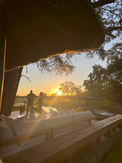 Here's a description of the image:
The image, apparently taken near sunset in Khwai, Botswana, is an outdoor scene featuring two silhouetted people on a wooden platform or deck. The sun is positioned on the horizon, creating a strong backlighting effect. Trees frame the upper portion of the image. The scene also includes a structure, possibly a building or the edge of a deck, on the right side, with cushions visible on seating. A rougher, more textured structure is on the left side of the frame. The silhouettes of the two people suggest one is standing with bent over and the other one has their hands on their hips. The environment appears to be natural, possibly near a river or field. The scene is strongly lit by the setting sun.