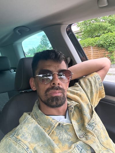A man is depicted relaxing inside a car, looking directly at the viewer. He is lying down in a passenger seat, with his head positioned on the left side of the frame and his body extending to the right. One arm is bent, with his hand resting behind his head, indicating a relaxed posture. He wears dark, round sunglasses with gold frames, which reflect the trees and sky outside. The man has short dark hair and a dark, neatly trimmed beard.
He is dressed in a patterned jacket, appearing to be denim-like, with a design in shades of yellow, blue, and grey. A white t-shirt is visible underneath the jacket. The car's interior is visible, featuring a grey fabric headliner, a dark grey headrest, and part of the seat.
Through the car's open sunroof or window, lush green foliage from trees is prominently displayed, suggesting a vibrant outdoor environment. A wooden fence or lattice structure is also discernible in the background outside the vehicle. The scene is brightly lit, indicating it is daytime. The combination of green trees and a fence implies a suburban or residential setting, consistent with the provided location context of Oberasbach, Germany. No visible text is present in the image.