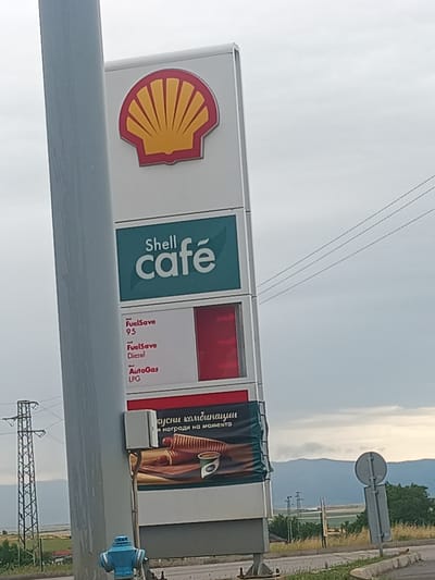 The image shows a Shell gas station sign in the city of Mogila, Bulgaria. The sign displays the Shell logo, the words "Shell café", and prices for FuelSave 95, FuelSave Diesel, and AutoGas LPG. Below the prices is a banner advertising a promotion with an image of pastries and a coffee cup. In the background, there are power lines, a power pylon, agricultural fields, and a mountain range under a cloudy sky. A blue fire hydrant is visible in the foreground.
