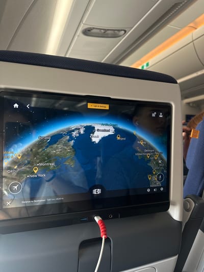 An in-flight entertainment screen displays a map of the Earth with flight path information. The screen shows locations such as Canada, Greenland, Iceland, Norway, the United Kingdom, Germany, France, and Spain. Notably, MUC is marked on the map, representing Munich Airport, which is located near the city of Freising, Germany. The screen also indicates a distance to destination of 7287 km or 4528 miles. A person's reflection is visible on the screen, wearing earbuds. To the right of the screen, a person wearing a blue shirt with an airline logo is partially visible. The background shows the interior of an airplane cabin with overhead lights and seats. A red and white cable is plugged into the bottom of the screen.