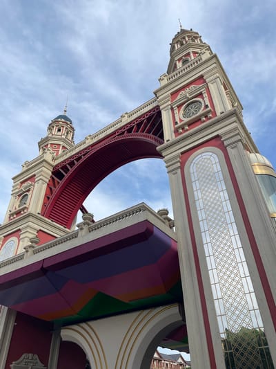 The photograph captures a low-angle view of a red and cream-colored bridge structure with ornate towers. The bridge features a prominent red archway with a latticed underside. The towers are decorated with recessed windows and detailed molding. Below the bridge, a colorful, geometric canopy is visible, displaying shades of purple, pink, orange, and green. In the background, a cloudy sky with blue patches is present. To the right of the main structure, a glass dome with a gold accent is attached to one of the towers. The reflection in the window panels reveals a patterned geometric design and greenery. The scene appears to be part of an architectural landmark in Nghĩa Trụ Commune, Vietnam, though no people or specific actions are depicted.