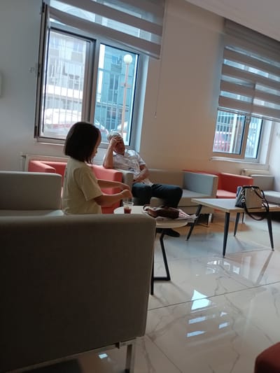 This media file shows a young girl and an older man in a waiting room. The girl is seated on a light gray couch on the left, facing away from the camera and slightly to the right. She has dark, shoulder-length hair and is wearing a light-colored t-shirt. She is holding a clear plastic cup with a red liquid and a stirring stick in her hands.

The older man is seated on a light gray couch on the right, facing the girl. He has gray hair and is wearing a light-colored patterned short-sleeved shirt and dark pants. He has his right hand on his forehead, suggesting he may be unwell or tired.

The waiting room has large windows with horizontal blinds that are partially open, allowing natural light to filter in. The room appears to be modern, with clean lines and a tiled floor that reflects the light. There are other seating areas visible, including a bright coral-colored couch. A light-colored coffee table is positioned between the two main couches, with a black handbag and some papers on it. A smaller, round table is near the girl, with her cup on it.

The overall impression is one of a calm, potentially clinical or administrative waiting area. The time of day appears to be daytime, given the natural light. The location is indicated as Ankara, Turkey. No text is visible in the image.
