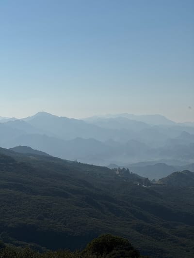 A panoramic landscape of the Unincorporated Santa Monica Mountains, United States of America, is depicted under a clear, pale blue sky. The foreground consists of densely vegetated, dark green hillsides. In the mid-distance, on a prominent ridge to the right, a small cluster of structures and associated trees are visible, indicative of limited habitation or infrastructure. Further into the hazy midground, a distinct communication tower or similar vertical structure is present atop a distant peak to the left. Beyond these, multiple successive ranges of mountains recede into the far distance, exhibiting strong atmospheric perspective as they fade into increasingly lighter shades of blue and grey due to significant haze. No human figures or discernible activities are present.