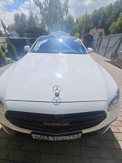 A white sedan is parked on a paved driveway in front of a house. The car has a Mercedes-Benz logo on the hood. The license plate is visible and reads "0031CC 797 RUS". In the background, there are trees, a fence, and a house with a red roof. The sky is blue with some clouds, and the sun is shining brightly, causing lens flare on the car's hood and windshield. The location is identified as Лужники Снт, Russia.