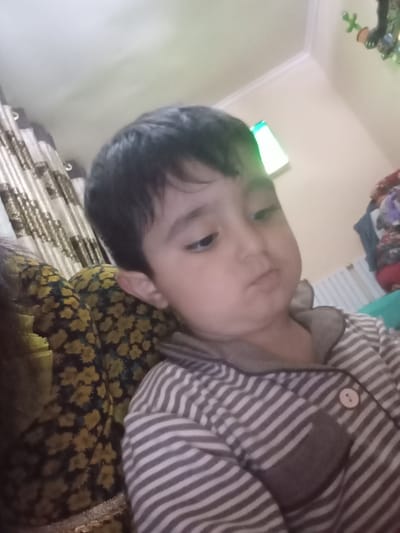 A young child with dark hair is pictured indoors. The child is wearing a gray and white striped garment and appears to be sitting next to a person wearing a dark green and yellow floral print top. The background includes a curtain, a white wall with a light fixture and a decorative item on the ceiling. There is no indication of the image being taken in Galaosiyo, Uzbekistan.