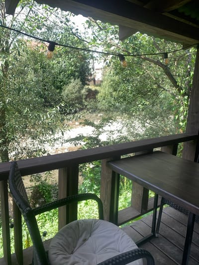 The image displays a view from a wooden balcony overlooking a river in Yasinia, Ukraine. In the foreground, a dark wicker chair with a white cushion is partially visible on the left, and a dark wooden table is partially visible on the right, both resting on a wooden plank floor. A dark wooden railing, supported by vertical posts, extends horizontally across the frame. Above, a string of Edison-style light bulbs is suspended. Beyond the railing, a shallow river with a light-colored, rocky bed flows through a landscape dominated by dense green trees and bushes. Indistinct structures, possibly buildings, are faintly visible through the foliage in the distant background. The scene is illuminated by daylight, and no individuals are present.