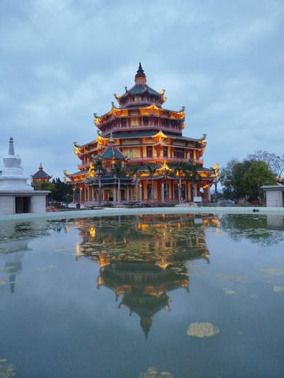 The image depicts a large, multi-tiered pagoda in Phú An, Vietnam, at dusk. The pagoda, illuminated with yellow lights, is reflected in a still body of water in the foreground. The structure is predominantly red and orange with green accents on the roof. The surrounding environment includes a white stupa-like structure to the left, other trees, and buildings in the background. The sky is overcast, creating a soft, diffused light.