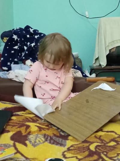 A young child with blonde hair is sitting on a colorful patterned rug and playing with a piece of white paper and a wooden board. The child is wearing a pink t-shirt with a cherry pattern. In the background, there is a blue blanket with white stars, and some folded clothes. The wall behind the child is painted a light blue-green color. A wooden shelf with some items on it is visible to the right. The overall scene suggests a casual indoor setting, possibly a bedroom or play area in a home in Kitskany, Moldova.