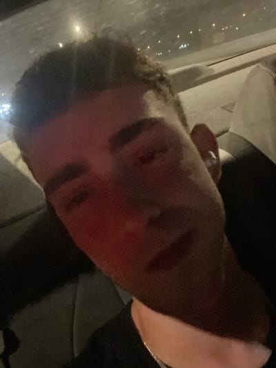 A young adult male is depicted in a close-up, dimly lit image, seated in the rear interior of a vehicle in Lod, Israel. The subject's face, primarily the left side, is illuminated by a distinct red light source, while the right side remains in shadow. He possesses dark, curly hair and is attired in a dark-colored shirt, complemented by a silver chain necklace. A white earbud is inserted into his right ear. The background reveals blurred points of light through the vehicle's window, indicative of an external urban environment at night. The visible vehicle upholstery is dark, likely gray or black.
