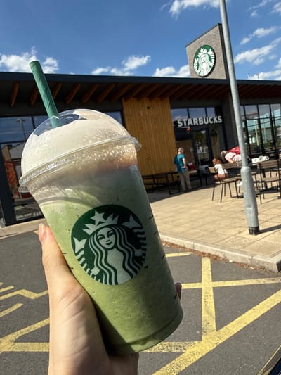 The image shows a Starbucks green tea frappuccino being held in front of a Starbucks store. The location is likely in Gedling, United Kingdom, based on available information. The Starbucks store is visible in the background with customers seated outside. The sky is blue with some clouds. The cup is a tall size, with a green paper straw and a layer of white foam at the top. A hand is holding the drink. Yellow lines painted on the pavement are visible in the foreground.