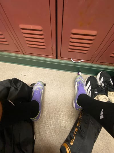 The image displays an indoor scene, likely a locker room or hallway, in the Town of Colonie, United States. The perspective is low, focusing on the floor and the base of a locker wall.

In the foreground, the lower legs and feet of two individuals are visible, resting on a light-colored, textured floor. On the left, a foot is clad in a purple and light green athletic shoe/cleat, paired with a black compression sock. A black athletic bag rests beside it. On the right, another foot wears an identical purple and light green athletic shoe/cleat with a black compression sock. Next to this foot, a pair of black Adidas sneakers with white stripes and light-colored socks are positioned. A black and orange bag, labeled "ELITE SPORT GOALKEEPING," is partially visible near the right foot. A small purple, cylindrical object lies on the floor between the purple shoes.

The background consists of a row of reddish-brown metal lockers, each featuring horizontal ventilation slats near the bottom. A dark green baseboard separates the lockers from the floor. A thin white wire or cord is present on the floor directly in front of the lockers.