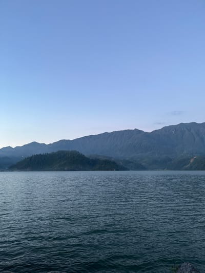 The image depicts a serene, expansive body of water in the city of Merodot, Indonesia. The water's surface is calm, with subtle ripples reflecting the soft, early morning or late evening light. Beyond the water, a range of lush green mountains rises, their slopes covered in dense vegetation. The sky above is a clear, pale blue, transitioning to a lighter hue towards the horizon. The overall scene conveys a sense of tranquility and natural beauty.