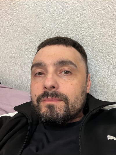A close-up headshot of a man with short, dark hair and a dark beard. He has light skin, brown eyes, and is wearing a black jacket. The background is a textured white wall. The image provides no information to suggest a location in the city of Kovel, Ukraine.