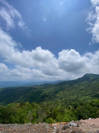 This image captures a panoramic view of lush, green mountains under a bright blue sky dotted with fluffy white clouds. The foreground features a rocky, disturbed area with some scattered debris, suggesting construction or land clearing. The rolling hills, covered in dense vegetation, extend into the distance, fading into a hazy, atmospheric perspective. The scene evokes a sense of natural beauty and vastness. The image is described as being taken in Nam Hải, Vietnam.