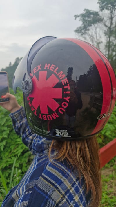 In Samoeng Tai, Thailand, a person wearing a blue and yellow plaid shirt is captured from the back, posing for a photograph with a smartphone. They are wearing a black and red striped motorcycle helmet adorned with a red starburst logo and the text "VEX HELMET! YOU MUST TAKE". The helmet has a clear visor. The background consists of lush green vegetation and a cloudy sky, indicating an outdoor setting. The person's hair, a light brown color, is visible beneath the helmet.