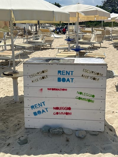 The image depicts a beachfront scene in Costa Rei, Italy. A white wooden kiosk, likely a rental booth, is positioned on the sand. The kiosk is adorned with stenciled text in various colors, advertising services such as "RENT BOAT," "ESCURSIONI ALLE ISOLE" (Excursions to the Islands), "INFORMAZIONI" (Information), and "NOLEGGIO GOMMONI" (Inflatable Boat Rental). In the background, rows of empty lounge chairs and umbrellas are set up on the sandy beach, suggesting a tourist area. Beyond the beach, vegetation and a cloudy sky are visible. The overall impression is that of a service provider on a tourist beach, ready to offer rentals and information to visitors.