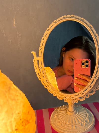 In the reflection of an ornate oval mirror, a young person is captured in a selfie. They are holding a pink phone with a polka-dot case, which has a triple camera system. The person's hair is dark brown, and they are wearing a pink long-sleeved shirt. Their gaze is directed at the camera, with a hint of a smile. The mirror is framed by a decorative, gold-colored metalwork that has a vintage aesthetic.
The background in the mirror's reflection is a deep blue. To the right of the mirror, a cream-colored lamp base with intricate detailing is visible. A pink and white striped surface is also partially visible in the background. The lighting suggests an indoor setting, possibly a room with a warm light source. The overall mood appears casual and personal, typical of a selfie taken by a young individual. The image does not contain any visible text.
