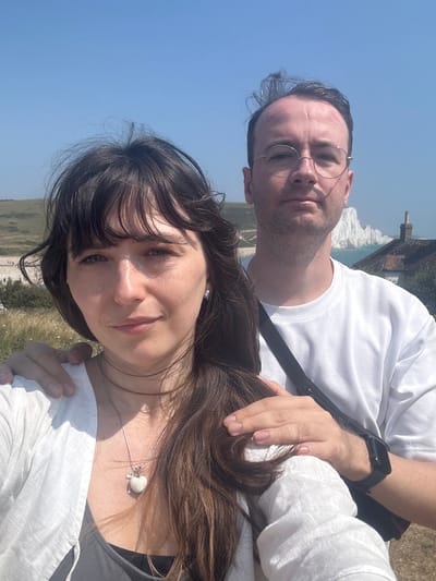A selfie taken outdoors shows a man and a woman standing close together with a scenic backdrop. The woman is in the foreground, facing the camera and smiling slightly. She has dark, long hair that is partially covering her face and is wearing a white top and a necklace with a heart pendant. The man is behind her, also looking at the camera, wearing glasses and a white t-shirt. His arm is around her shoulder. In the background, there are rolling green hills, a body of water, and white cliffs, consistent with the location of Seaford, United Kingdom, specifically near the Seven Sisters chalk cliffs. The sky is clear and blue.