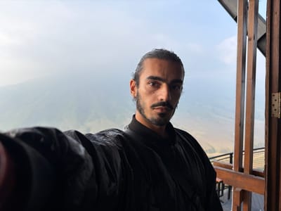 A selfie taken by a man with dark hair pulled back, a mustache, and a goatee. He is wearing a black bomber jacket and a black shirt. The man is standing on a wooden balcony with a railing overlooking a misty valley. The location is identified as Ngadas, Indonesia.