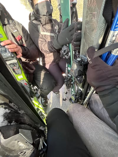 The image depicts the interior of a ski lift gondola or cable car cabin in Villard-sur-Doron, France, occupied by at least four individuals in full ski attire, along with their equipment. The perspective is from a seated or lower vantage point, looking across and slightly down.
Essential details:
* **People:** Four individuals are visible.
* One person on the far left wears a dark jacket, holds a neon green ski with black FX bindings, and has a dark strap with a red buckle visible. Their hands are gloved.
* Another person in the center wears a dark "THE NORTH FACE" jacket with two horizontal grey reflective stripes, dark pants, and a black "MON TEC" perforated neck gaiter covering their lower face. Black "WEDZE" branded gloves are visible on their hands, which are holding a green ski with black bindings. Black ski boots are present on their feet.
* A third person, on the right, wears dark pants and dark "Reusch" branded gloves, holding blue ski poles and skis with textured grey surfaces and black bindings.
* The fourth person is only partially visible in the foreground, showing legs in black and grey mottled ski pants, implying the camera operator or a companion.
* **Objects:** Multiple pairs of skis with various black bindings are prominent. Specific brands visible include "FX" on a neon green ski binding, "THE NORTH FACE" on a jacket, "MON TEC" on a neck gaiter, "WEDZE" on a glove, and "Reusch" on another glove. Blue ski poles are also present.
* **Actions:** The individuals appear to be seated or standing within the enclosed transport, holding their ski equipment, indicating transit within a ski resort.
* **Environment:** The setting is an enclosed cabin with a grey, textured floor. Bright sunlight illuminates the scene, suggesting an outdoor, daytime winter environment, likely with snow-covered slopes or trees visible outside the unpictured windows. Some snow or ice accumulation is visible on the far-left edge of the cabin's interior.