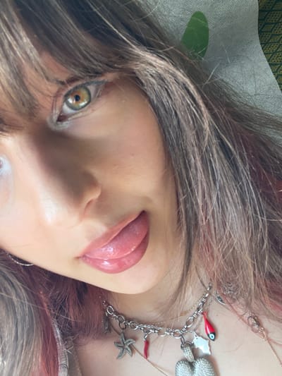 An extreme close-up photograph depicts a female individual in Tzafria, Israel, positioned at an upward angle. Her left eye, characterized by a green-hazel iris with a dark pupil and eyeliner, is visible at the top center. Brown hair with reddish-pink highlights frames the left side of her face and extends across the upper forehead as bangs. Her mouth is partially open, revealing a glossy, pink-toned tongue extended outwards. Multiple layered necklaces are visible below her chin, including a silver-toned choker adorned with various charms: a starfish, a red chili pepper, a fish, a white outline charm, and a large textured heart-shaped pendant. A subtle silver hoop earring is present on the left side of her face. The background consists of a light-colored, textured surface on the upper right, partially obscuring a dark green, patterned fabric.