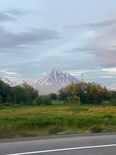 The image depicts a majestic snow-capped volcano dominating the background, its slopes crisscrossed with snowfields and shadows. In the middle ground, a line of trees separates the volcano from a grassy field in the foreground. A power line stretches across the scene, supported by a tall tower. The sky above is filled with soft, diffused clouds, suggesting either early morning or late evening light. The bottom of the image shows a blurred asphalt road with a white line, indicating that the photo was taken from a moving vehicle. The overall atmosphere is serene and natural, with no human presence visible. The location is identified as the city of Ketkino, Russia.