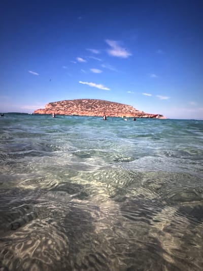 The image shows a vast expanse of clear, turquoise water in the foreground, with ripples and sunlight creating a dynamic pattern on the surface. In the mid-ground, several people are visible in the water, some appearing to be swimming or wading. Further in the distance, a large, rocky island or promontory dominates the horizon. This landmass is covered in reddish-brown vegetation and appears to be a prominent natural feature. The sky above is a vibrant blue, with a few wispy white clouds scattered across it. The overall scene depicts a lively beach or coastal area, with people enjoying recreational activities in a natural setting. The image does not contain any elements that would allow for identification of the specific location within the city of Porrentruy, Switzerland.