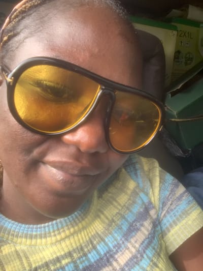 A close-up, head and shoulders, selfie-style photograph shows a dark-skinned woman wearing large, dark-rimmed sunglasses with yellow lenses. Her hair is pulled back in braids. She is wearing a brightly colored, striped sweater in shades of yellow, blue, and grey. In the background, a green box with labels can be seen. The image appears to be taken outdoors during the day. The location is identified as Assob Hausa, Nigeria.