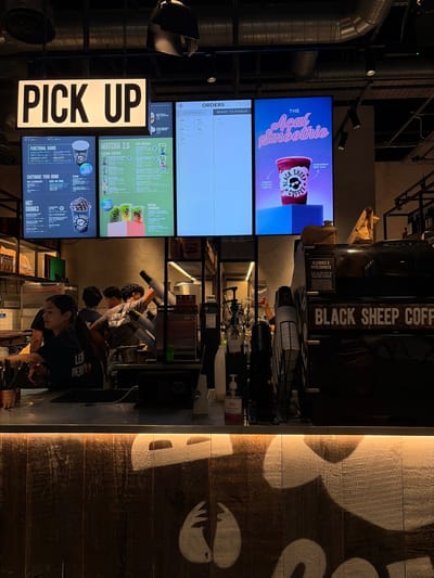 The image depicts the interior of a "Black Sheep Coffee" shop, a popular chain, likely located in Luton, United Kingdom, judging by the overall aesthetic and common presence of such establishments in UK high streets. The scene is captured from a slightly low angle, looking towards a counter area where staff are actively preparing orders.
Dominating the upper part of the frame are three large digital screens. The leftmost screen, illuminated with a bright white light and bold black text, displays "PICK UP," indicating a designated order collection point. To its right, a screen is divided into two sections: the left displays order numbers and status ("PREPARING...", "READY TO PICKUP"), with the time "09:59" visible. The rightmost screen features a vibrant advertisement for "THE Acai Smoothie" from "BLACK SHEEP COFFEE," showcasing a purple smoothie in a clear cup.
Below these screens, a menu board is visible, detailing various product categories: "FUNCTIONAL RANGE," "MATCHA 2.0," and "CUSTOMISE YOUR DRINK," along with "HOT DRINKS" such as "SIGNATURE HOT CHOCOLATE" and "TURMERIC LATTE."
The counter area itself is a hive of activity. Three young individuals, presumably staff members, are engaged in drink preparation. One person, wearing a black t-shirt with "LEAVE HER BEHIND" printed on it, is in the foreground, leaning on the counter and looking to the left. Behind them, two other individuals are working, one wearing a grey t-shirt and the other a darker shirt, obscured by equipment. Various coffee-making appliances, cups, dispensers, and other operational items are visible on the counter.
To the right of the counter, a large black coffee machine is prominent, bearing the "BLACK SHEEP COFFEE" logo. Above the coffee machine, a sign details "ALLERGIES & INTOLERANCES." The overall environment is dimly lit, with warm, industrial-style lighting emanating from above and along the front of the counter, highlighting the wooden texture of the counter's facade. The background suggests a modern, urban commercial space with exposed ductwork and ceiling fixtures.