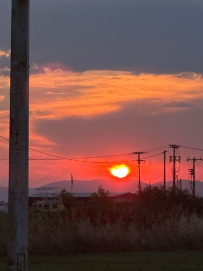 An outdoor, low-angle, vertical shot captures a sunset in Chaidefto, Greece. The sky transitions from dark gray at the top to vibrant orange and red hues closer to the horizon, illuminated by the setting sun. The sun itself appears as a bright, heart-shaped orb, emitting a warm glow. In the foreground, a weathered utility pole stands to the left, partially obscuring the view. Silhouetted against the colorful sky are distant mountains, power lines supported by utility poles, and the outlines of buildings and dense foliage. The overall scene is atmospheric and dramatic, dominated by the radiant sunset.