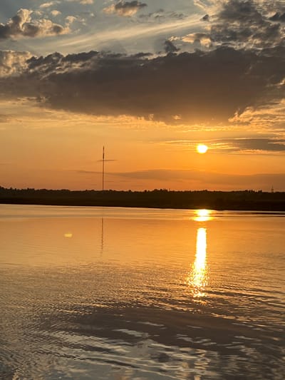 This is a vertical, eye-level, full shot of a sunset over a body of water, likely a lake or river in or near Nasva alevik, Estonia. The sky is a vibrant orange and yellow gradient, with streaks of grey and white clouds scattered throughout. The sun, a bright orange orb, is positioned in the upper right quadrant of the image, casting a brilliant reflection on the water's surface.
The water itself is calm, with gentle ripples disturbing its glassy surface. The reflection of the sun creates a shimmering, golden path extending from the horizon towards the viewer. In the distance, a dark, silhouetted tree line separates the water from the sky. A tall, thin tower, possibly a radio mast or communication tower, stands prominently against the sunset on the horizon, its reflection also visible in the water. No people or animals are visible in the scene. The overall impression is one of serene natural beauty, with the warm tones of the sunset dominating the composition.
