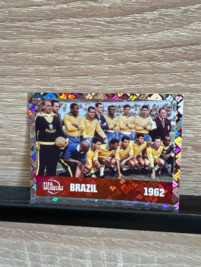 The image shows a rectangular trading card featuring a photograph of the Brazil national football team from 1962. The card is placed on a wooden surface with a visible grain pattern. The photograph depicts a group of men in football kits, some standing and some kneeling, posing for a picture. The card has holographic borders. Along the bottom edge of the card, there is text that reads "FIFA MUSEUM BRAZIL 1962". There are no discernible elements in the image that would suggest a location in Prisovo, Bulgaria, or any specific city. The scene captured in the photograph is a sports team photo taken outdoors, likely on a football field.