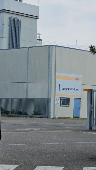The image shows the exterior of a light-colored industrial building with a gray metal fence in the foreground. A sign on the building, partially obscured by a fence post, displays the text "Göbel Getränke GmbH" and an arrow pointing upwards next to the words "Leergutabholung," which translates to "Empty Goods Collection." Below this sign, there is another smaller sign with a triangle indicating a warning and the text "Warenannahme," meaning "Goods Reception," next to a diagram of a pallet and a forklift. A blue door is visible on the building to the right of the signs. The scene appears to be in an industrial area with an asphalt road in front of the building. The sky is overcast. The image was taken in Neu-Ulm, Germany.