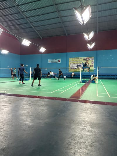 The image depicts an active indoor badminton court, captured from an elevated perspective looking down its length. Several male individuals are present, engaged in playing or practicing badminton. One person is seated in a high umpire's chair positioned next to the net.
The court features a vibrant green surface with crisp white boundary lines, bordered by a reddish-brown strip. The surrounding walls are a two-tone blue, with a lighter shade at the bottom and a darker blue above. The ceiling consists of corrugated metal sheeting supported by exposed beams, from which multiple powerful, rectangular floodlights hang, brightly illuminating the entire space.
On the blue wall, two banners are visible. A larger banner displays "SURYA KENCANA" alongside a crest-like logo and additional Indonesian text, likely indicating a club or event. Below it, a smaller red and white banner carries the message "TERIMA KASIH UNTUK TIDAK MERUSAK AREA LAPANG !!!", which translates to "THANK YOU FOR NOT DAMAGING THE COURT AREA !!!". A few water bottles are placed near the net. To the right of the court, a dark grey, textured concrete floor extends, possibly an adjacent pathway or viewing area. The artificial lighting suggests the activity is taking place indoors, suitable for any time of day or night, within a sports facility in Kecamatan Cisaat, Indonesia.