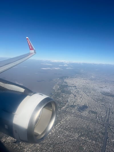 An aerial photograph captures a view from an aircraft, showcasing a segment of its port wing, red winglet with "JETSMART.COM" branding, and the forward section of the engine nacelle in the lower left foreground. The scene depicts a vast urban sprawl along the Río de la Plata, in the northern part of Greater Buenos Aires, Argentina, specifically over areas consistent with Martínez. The river, a wide expanse of brownish-grey water, occupies the left-mid portion of the frame, bordered by a coastline featuring green spaces and densely packed residential and commercial buildings laid out in a distinct grid pattern. The urban area extends inland, characterized by numerous white and grey structures forming a continuous settlement. The sky above is clear blue with a horizon line marked by scattered white clouds. No individuals or specific actions are discernible at this altitude.